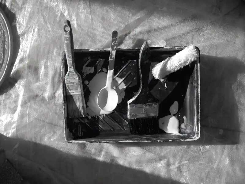Bac à peinture, pinceaux et rouleau posés sur une bâche, en noir et blanc